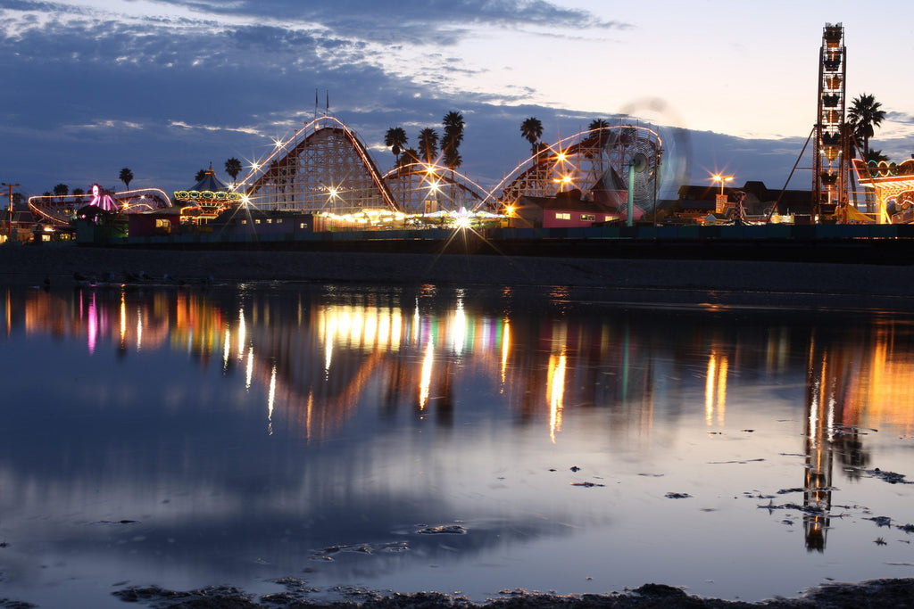 A seaside amusement park with a roller coaster and Ferris wheel reflected in still water at dusk