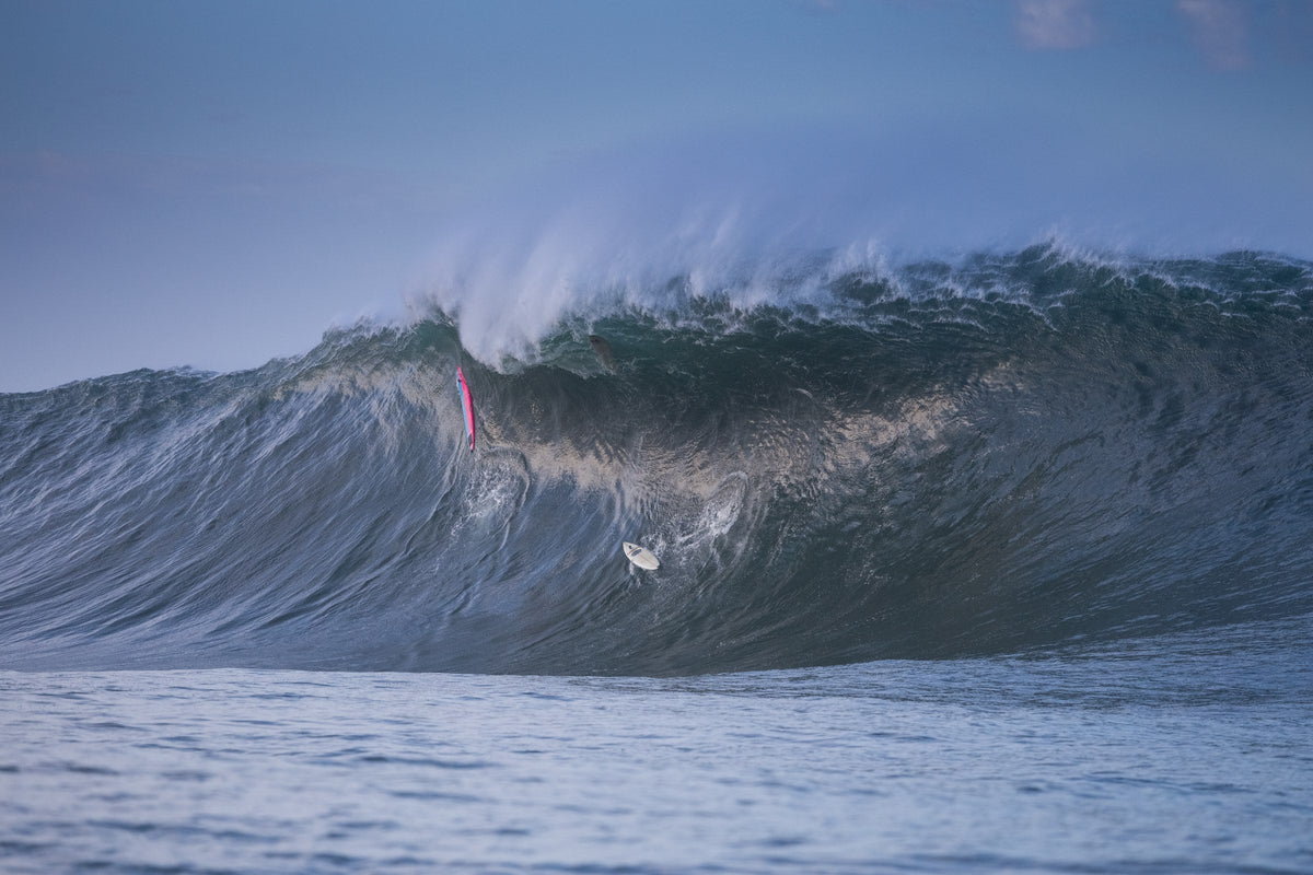 A big dark wave with spray blowing off the top and a pink surfboard on the face