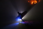 A silhouetted figure carrying a surfboard through beams of blue light on a dark beach with a warm glow on the rocks behind