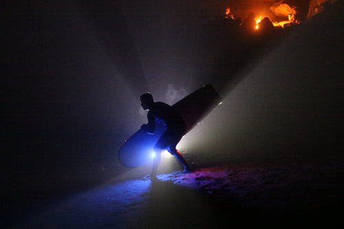 A silhouetted figure carrying a surfboard through beams of blue light on a dark beach with a warm glow on the rocks behind