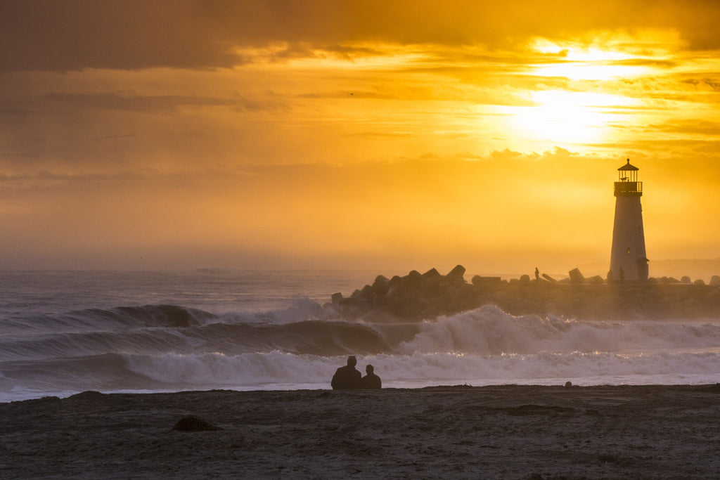 A lighthouse on a breakwater under a golden sunset with a couple watching from the shore
