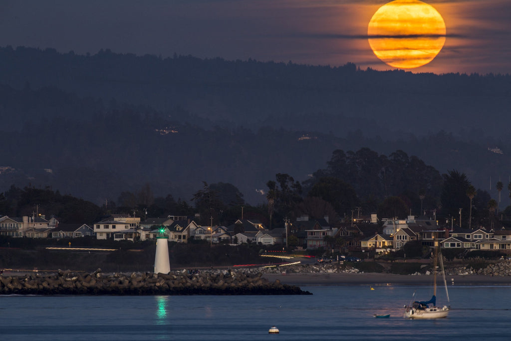 A huge orange full moon rising above a harbor town with a lighthouse and a sailboat