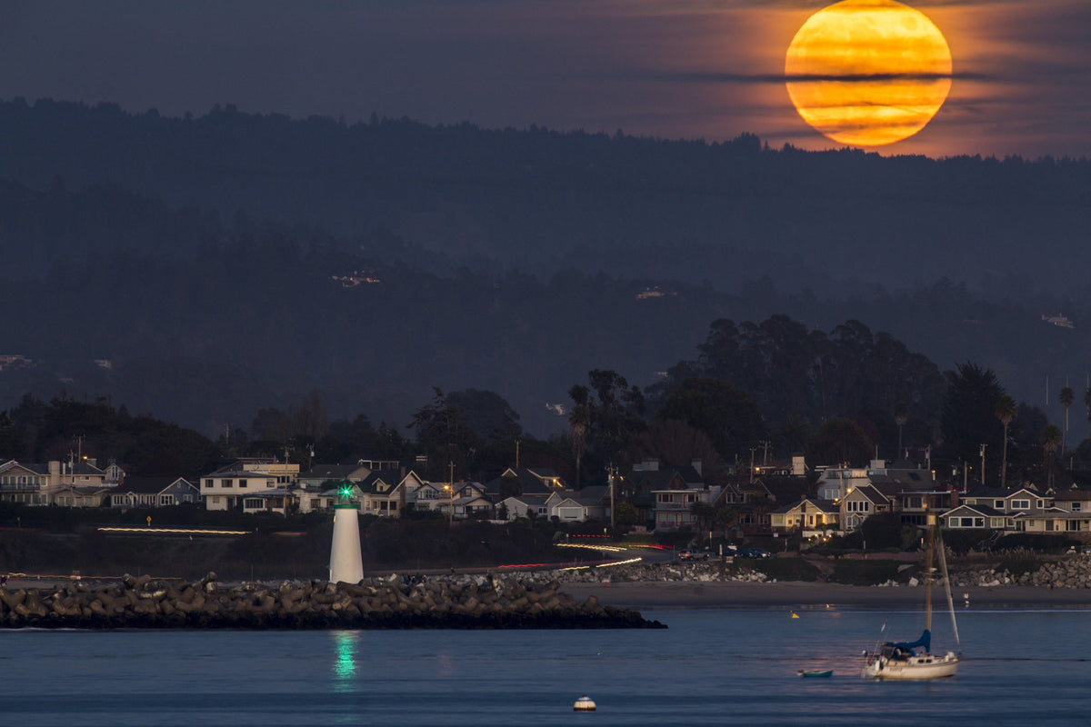 A huge orange full moon rising above a harbor town with a lighthouse and a sailboat