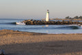A lighthouse at the end of a rock breakwater with a small wave peeling beside it and a surfer in the water