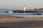 A lighthouse at the end of a rock breakwater with a small wave peeling beside it and a surfer in the water