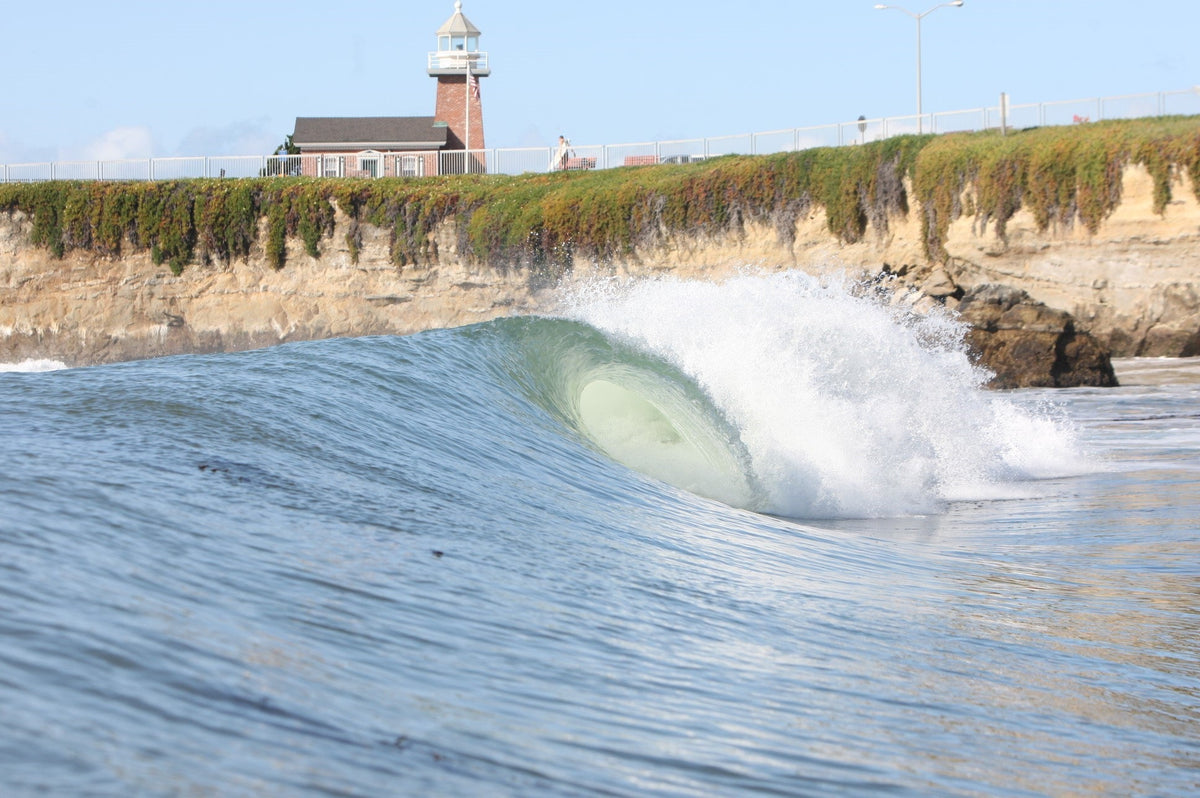 A glassy wave breaking along a cliff below a brick lighthouse