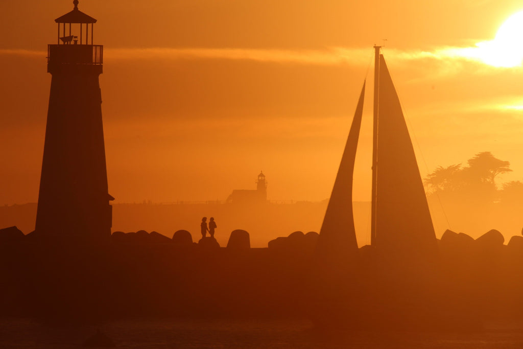A lighthouse and a sailboat sail in silhouette against an orange sunset