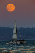 An orange full moon over a sailboat on the water at dusk