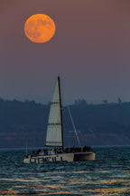 An orange full moon over a sailboat on the water at dusk