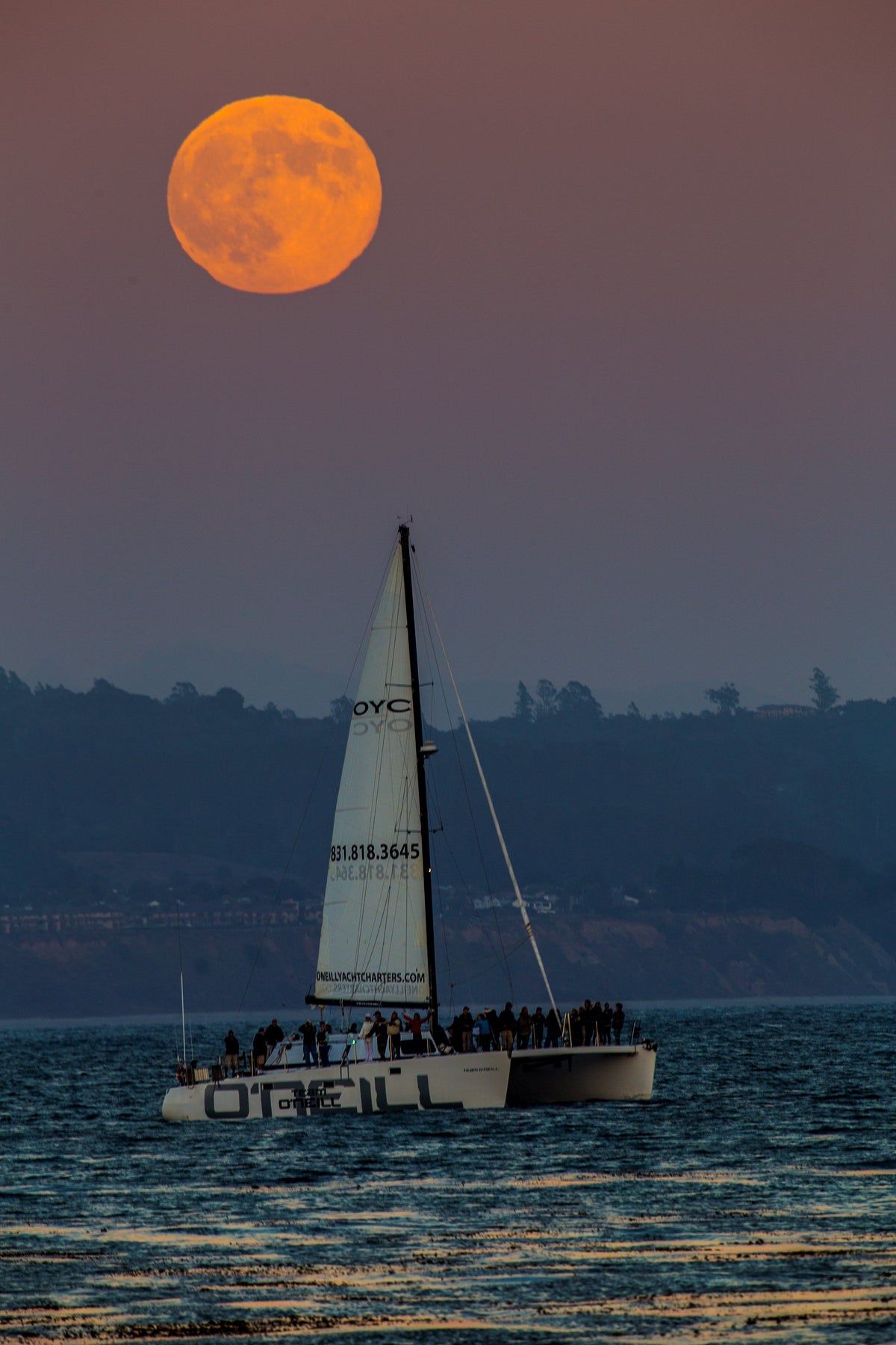 An orange full moon over a sailboat on the water at dusk