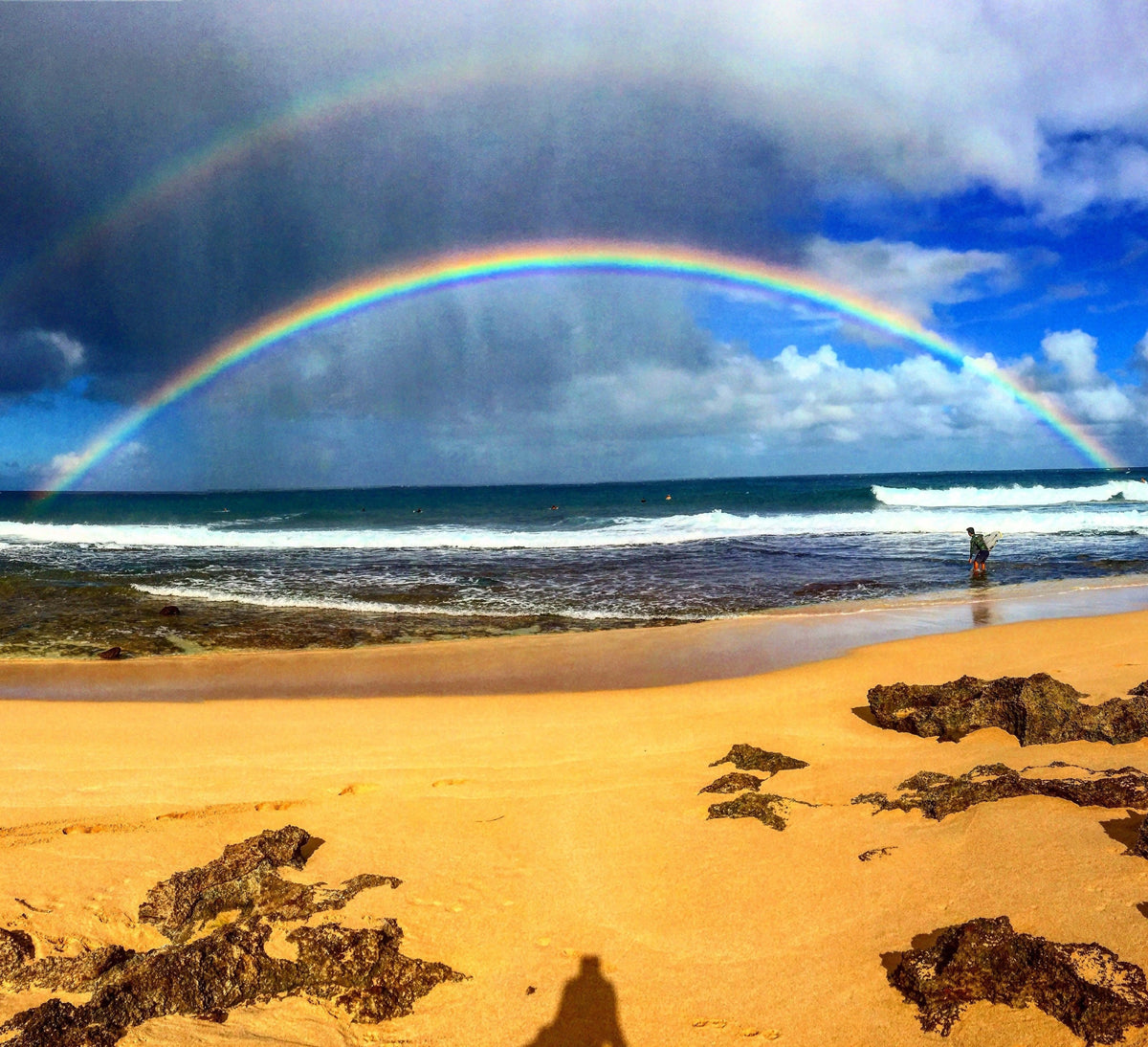 A rainbow over a gold sand beach with a lone person at the waterline