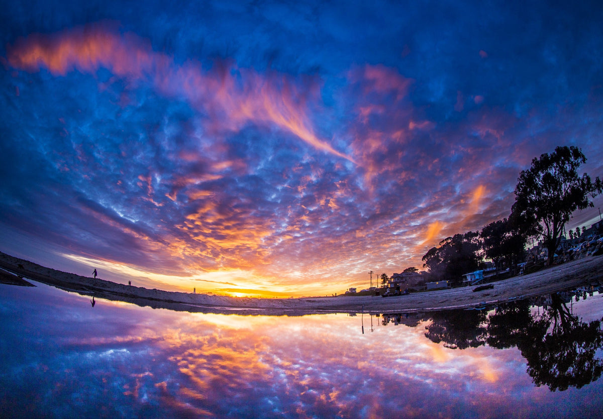 A sunset sky of purple and orange clouds reflected in a still puddle on the beach