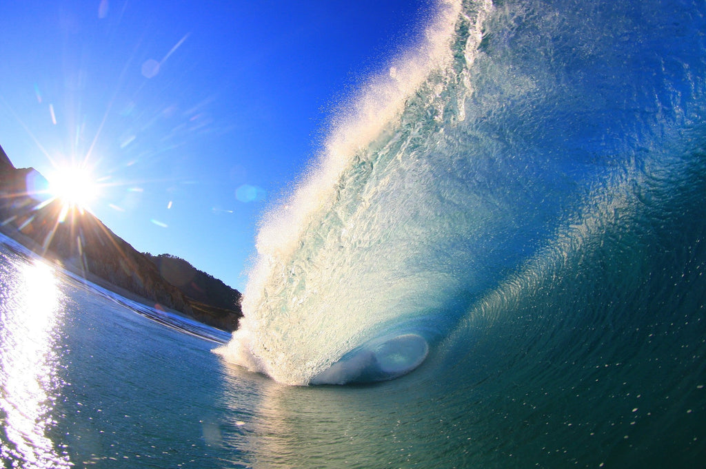 A bright blue wave lit by a sunburst over a rocky point
