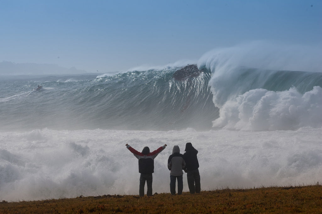 Three people with arms raised on a bluff facing an enormous wave and cloud of spray