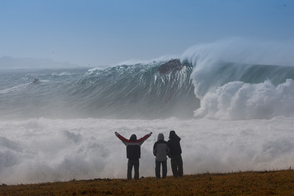 Three people with arms raised on a bluff facing an enormous wave and cloud of spray