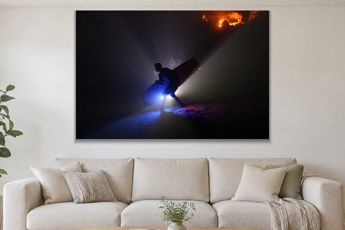 A silhouetted figure carrying a surfboard through beams of blue light on a dark beach with a warm glow on the rocks behind shown large on the wall of a lived-in room