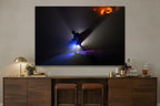 A silhouetted figure carrying a surfboard through beams of blue light on a dark beach with a warm glow on the rocks behind shown large on the wall of a lived-in room