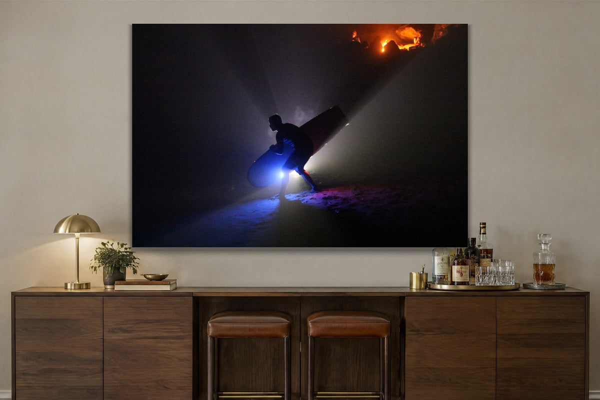 A silhouetted figure carrying a surfboard through beams of blue light on a dark beach with a warm glow on the rocks behind shown large on the wall of a lived-in room