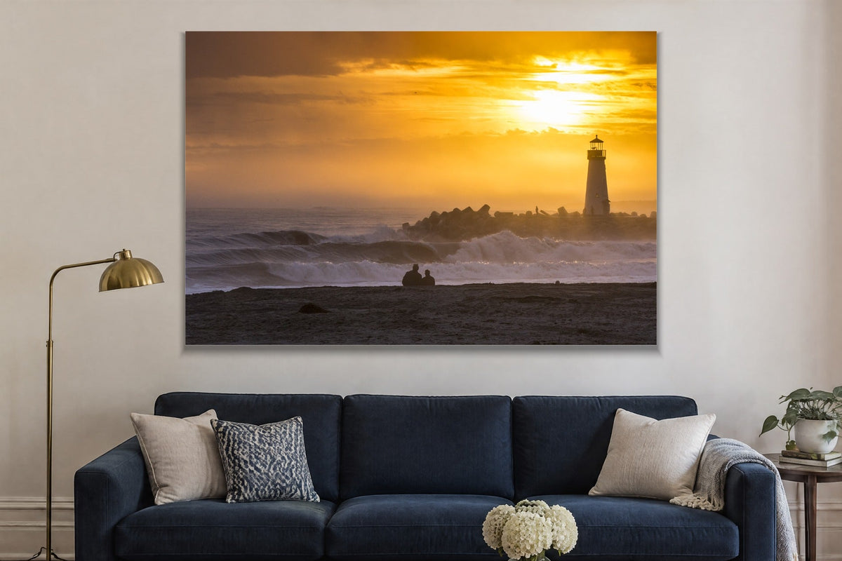 A lighthouse on a breakwater under a golden sunset with a couple watching from the shore shown large on the wall of a lived-in room