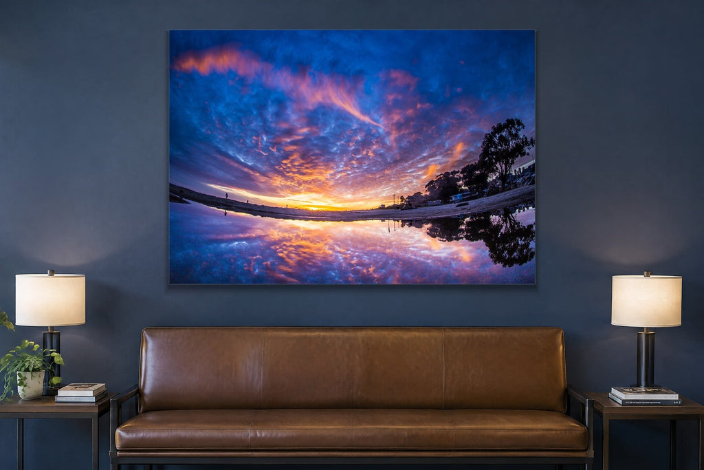 A sunset sky of purple and orange clouds reflected in a still puddle on the beach shown large on the wall of a lived-in room