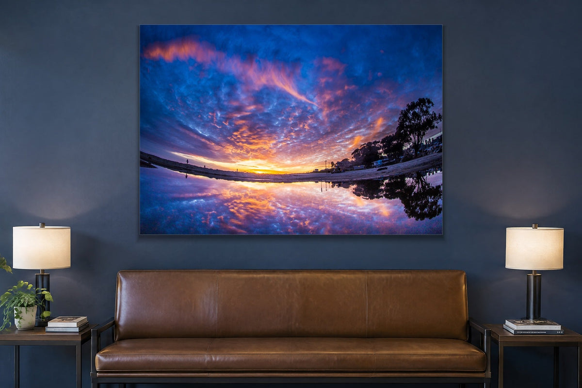 A sunset sky of purple and orange clouds reflected in a still puddle on the beach shown large on the wall of a lived-in room