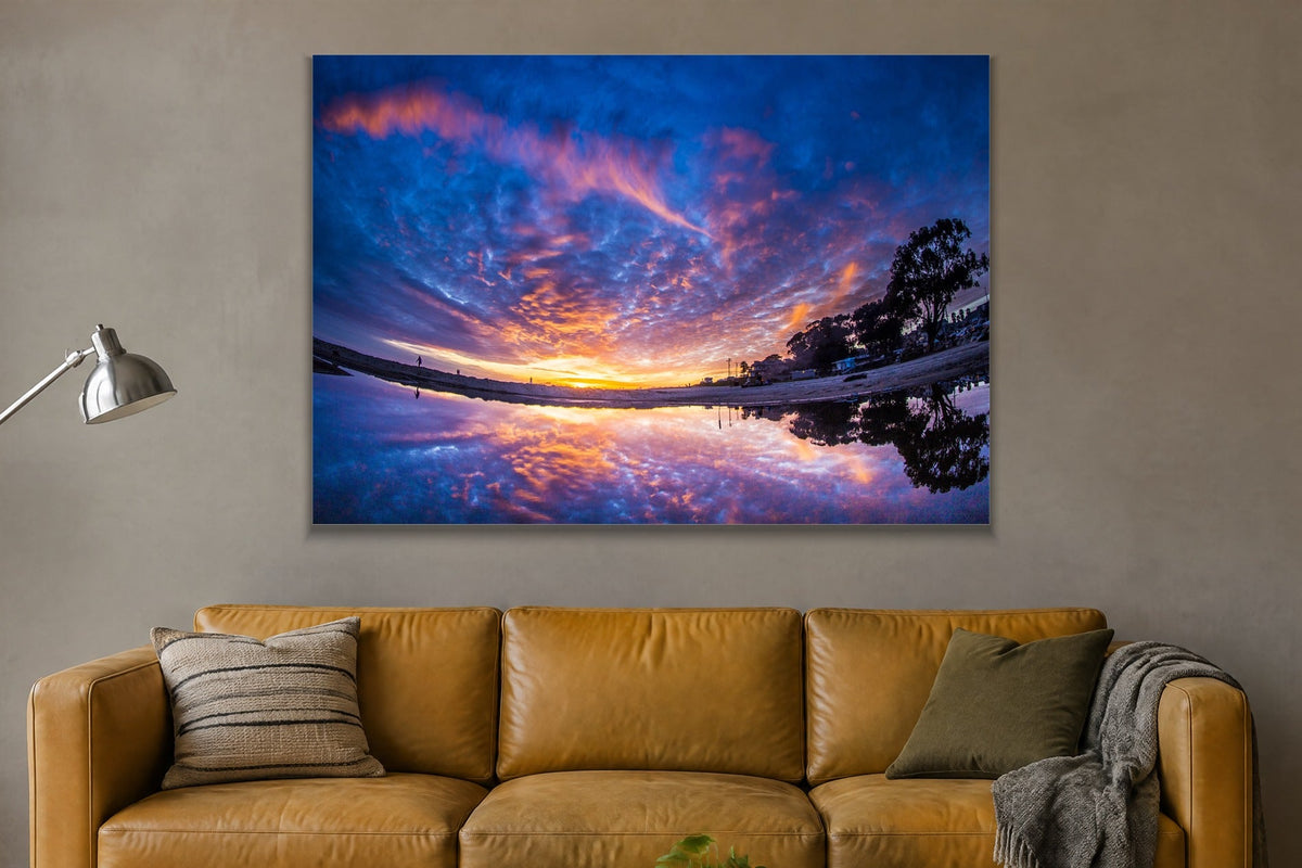 A sunset sky of purple and orange clouds reflected in a still puddle on the beach shown large on the wall of a lived-in room