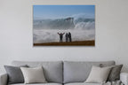 Three people with arms raised on a bluff facing an enormous wave and cloud of spray shown large on the wall of a lived-in room