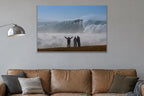 Three people with arms raised on a bluff facing an enormous wave and cloud of spray shown large on the wall of a lived-in room