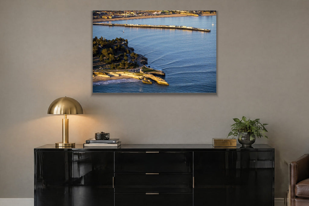 An aerial view of a long wharf and rocky point in blue water at golden hour shown large on the wall of a lived-in room